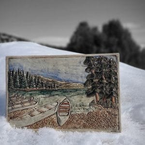 A ceramic art tile with a mountain scene with canoes waiting on the shore line. 